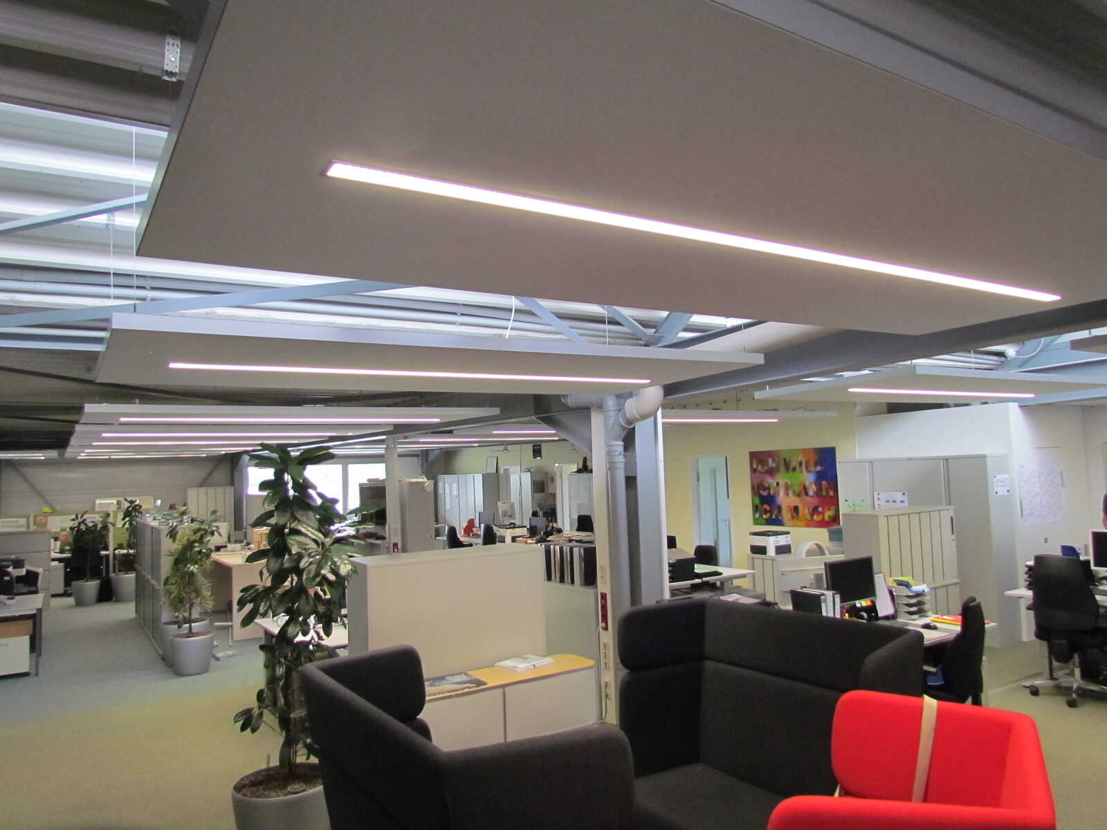 Großraumbüro mit großflächigen Deckensegeln und Lichtbändern über Arbeitsplätzen und Lounge-Zone