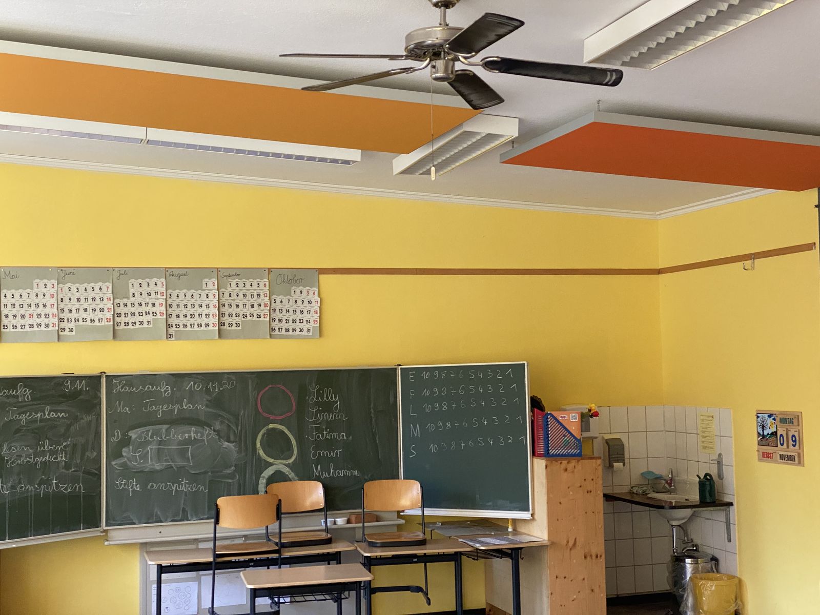 Klassenzimmer mit orangefarbenen Deckenabsorbern über Tafel und Pultreihen