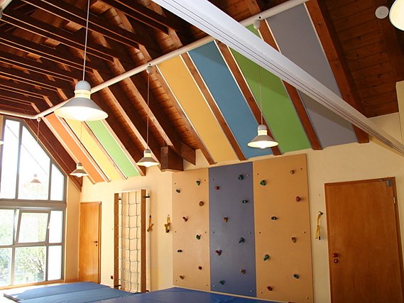 Farbige Akustikpaneele zwischen den Holzbindern einer Turnhallendecke, darunter eine Kletterwand